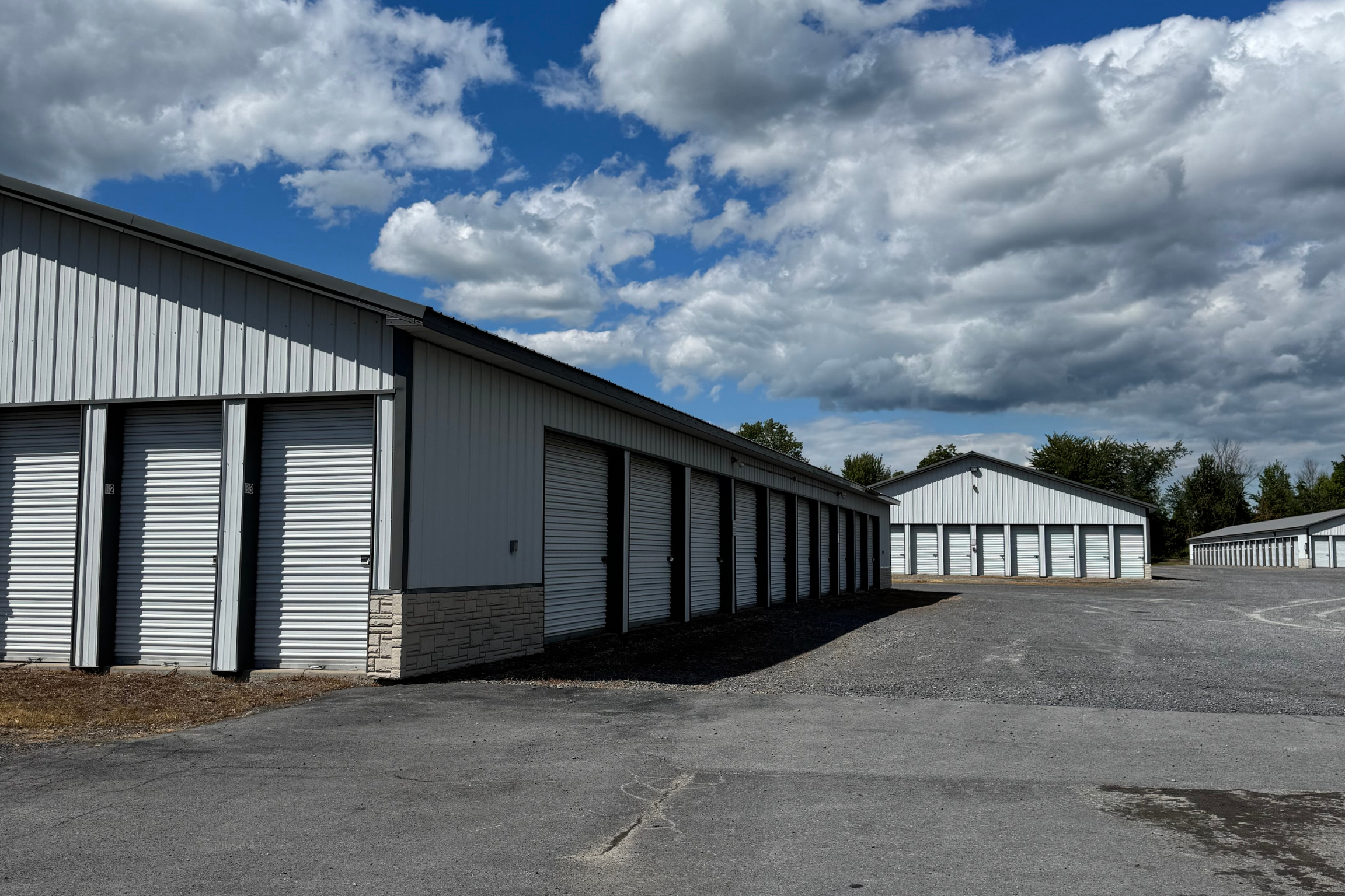 A row of self-storage units with white roll-up doors under a blue sky with scattered clouds. The paved driveway leads between multiple storage buildings.
